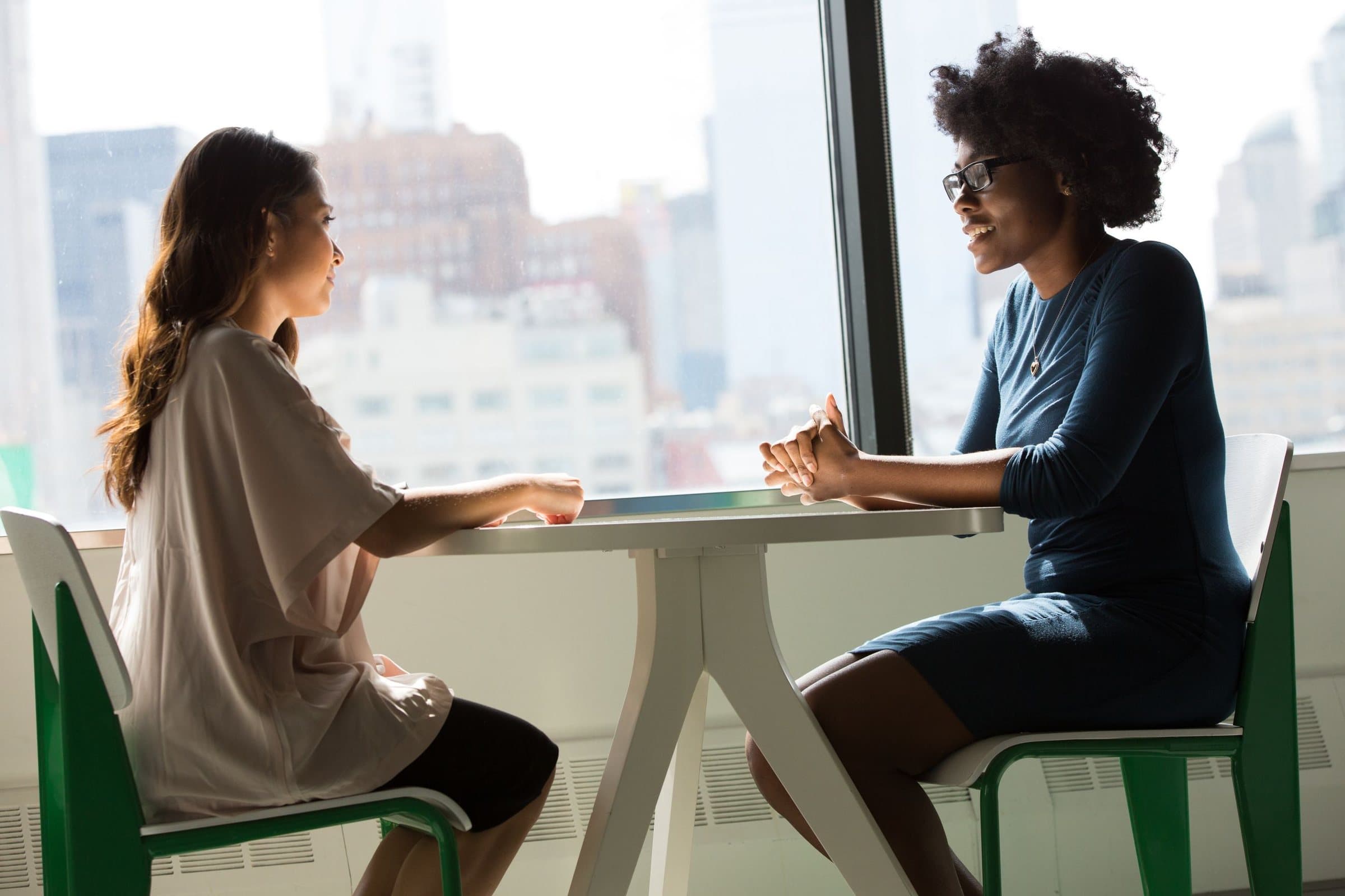Two women talking across a table by a window, with the city behind them